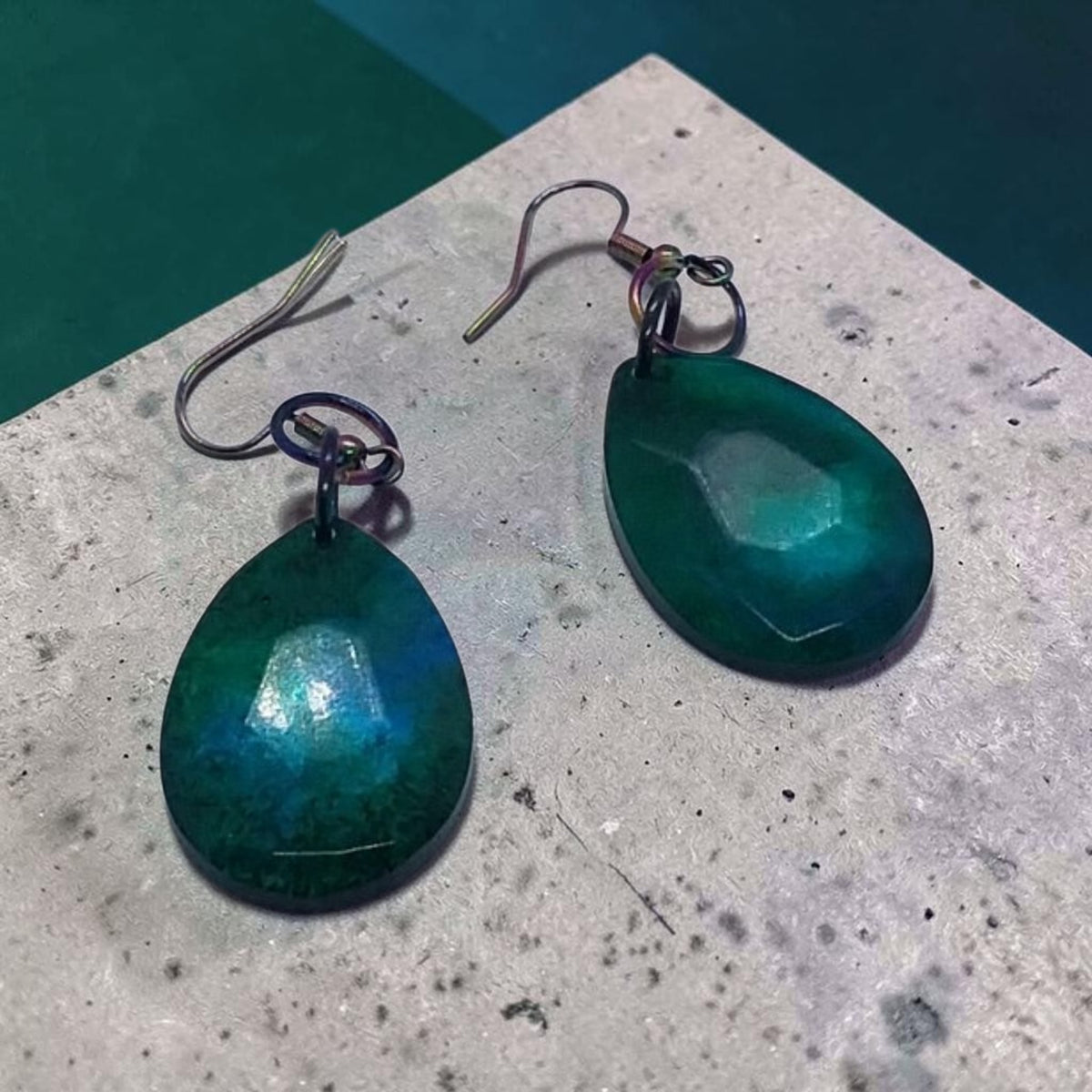 Artymunk3y teardrop earrings in teal and emerald resin with faceted surfaces and iridescent oil-slick metal hooks.