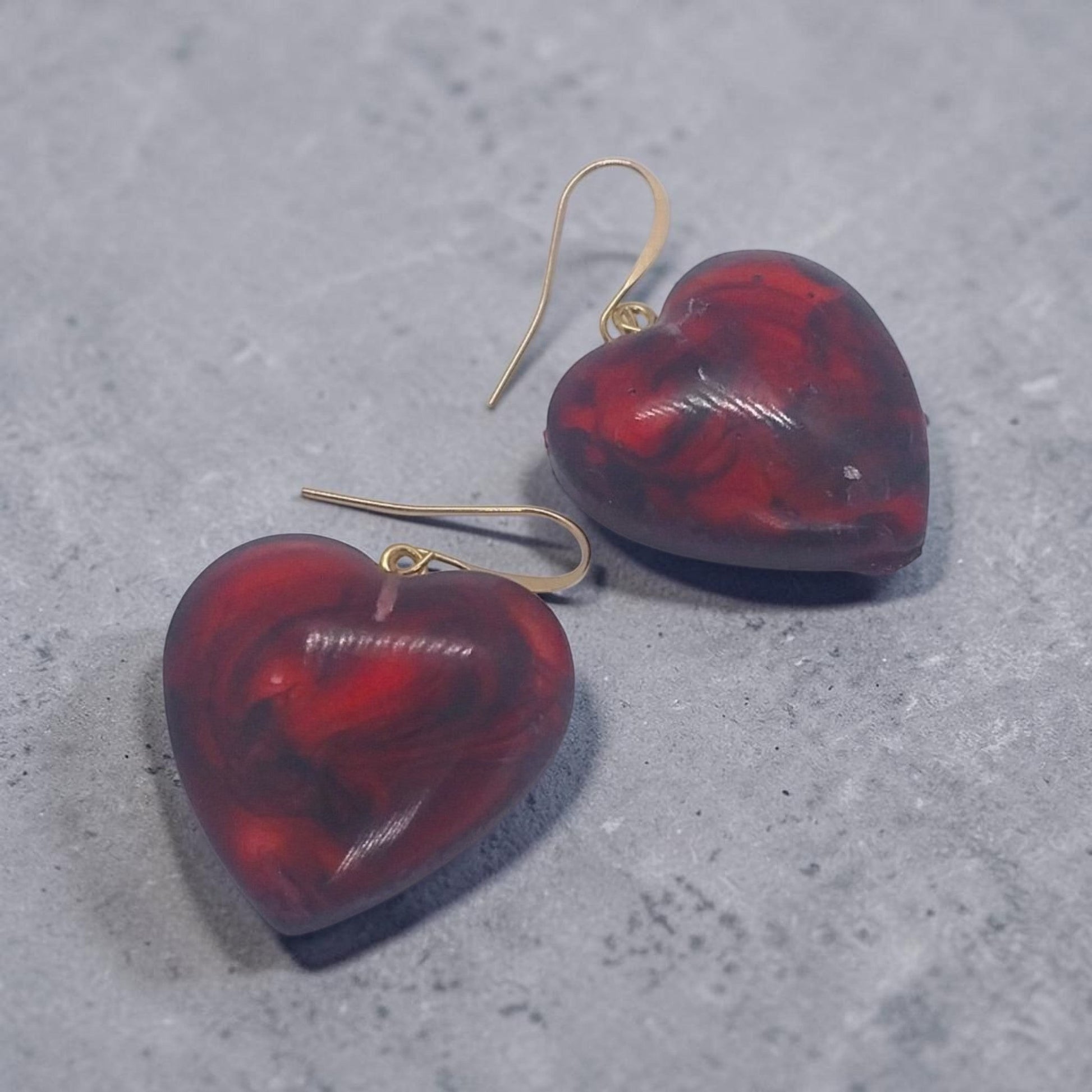Artymunk3y heart-shaped dangle earrings in marbled red and black resin with gold-tone hooks on a grey surface.