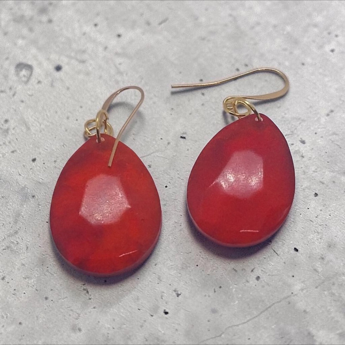 Artymunk3y faceted red teardrop dangle earrings with gold-tone hooks on a grey textured surface.