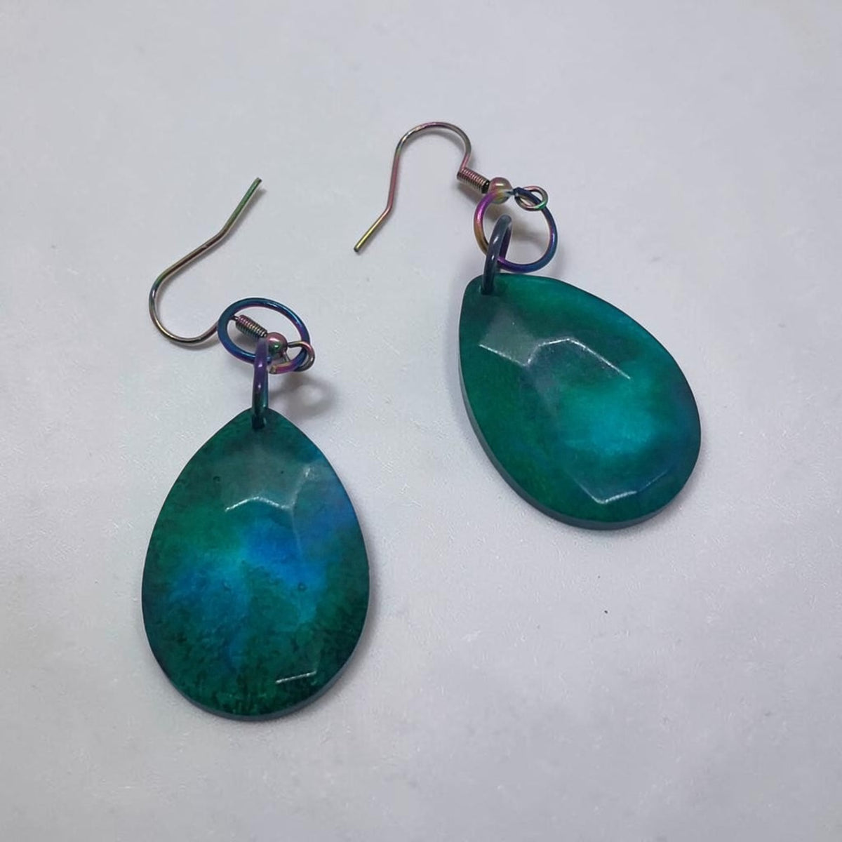 Artymunk3y teardrop dangle earrings in teal and emerald resin with faceted surfaces and iridescent oil-slick hooks.